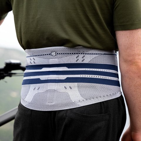 Mann mit grünem T-Shirt trägt eine graue Rückenbandage mit blauen Streifen, von hinten fotografiert beim Fahrradfahren mit Blick auf eine bergige Landschaft.