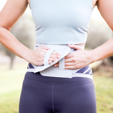 Frau im Sportoutfit befestigt eine graue Rückenbandage mit Klettverschluss um ihren unteren Rücken, im Freien bei Tageslicht.