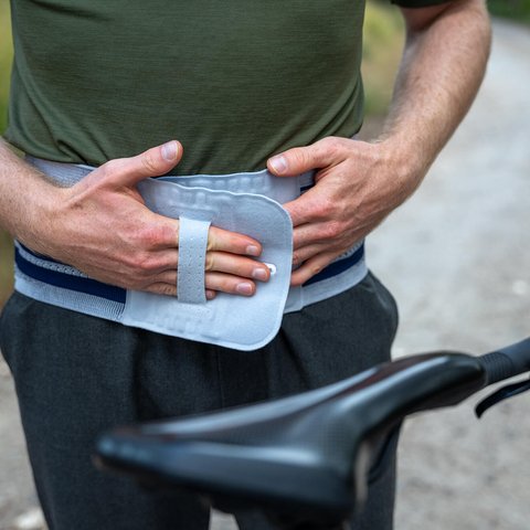 Mann mit grünem T-Shirt fixiert den Klettverschluss einer Rückenbandage an seinem Bauch, neben einem Fahrrad auf einem Waldweg.