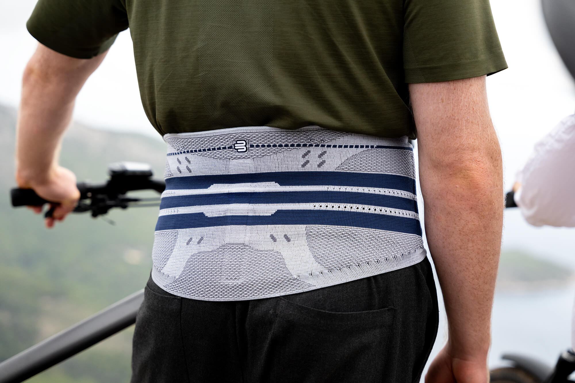 Mann mit grünem T-Shirt trägt eine graue Rückenbandage mit blauen Streifen, von hinten fotografiert beim Fahrradfahren mit Blick auf eine bergige Landschaft.
