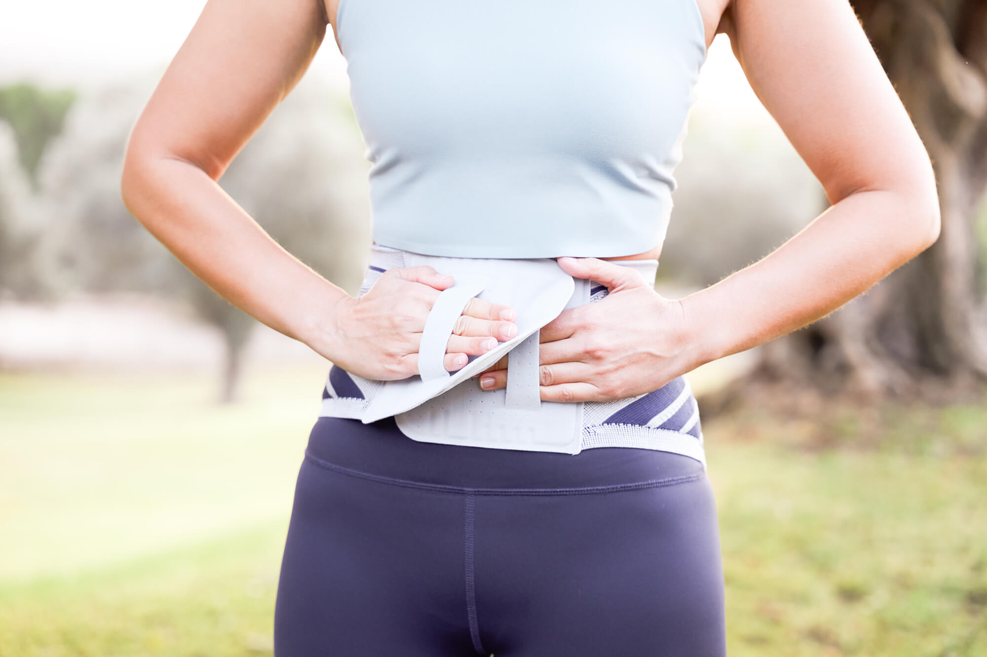 Frau im Sportoutfit befestigt eine graue Rückenbandage mit Klettverschluss um ihren unteren Rücken, im Freien bei Tageslicht.